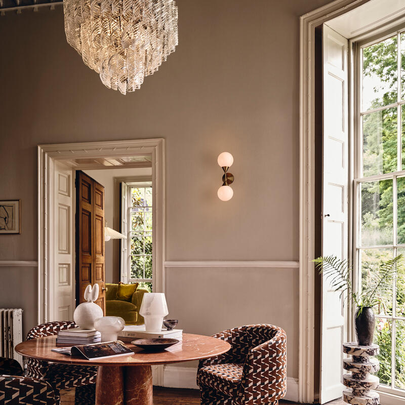 A round wooden dining table with assorted books, sculptures, and a lamp sits in a stylish room featuring patterned chairs, a large window with a garden view, and a chandelier above.