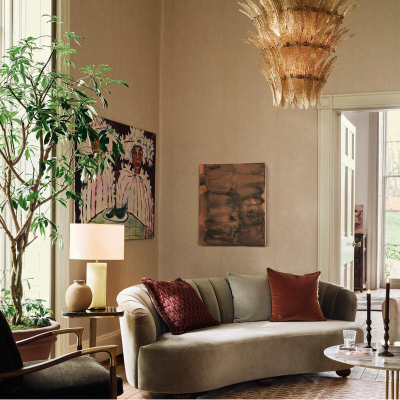 An elegant living room features a curved sofa with contrasting pillows, a tall plant, an ornate chandelier, and artwork on beige walls, all bathed in natural light from large windows.