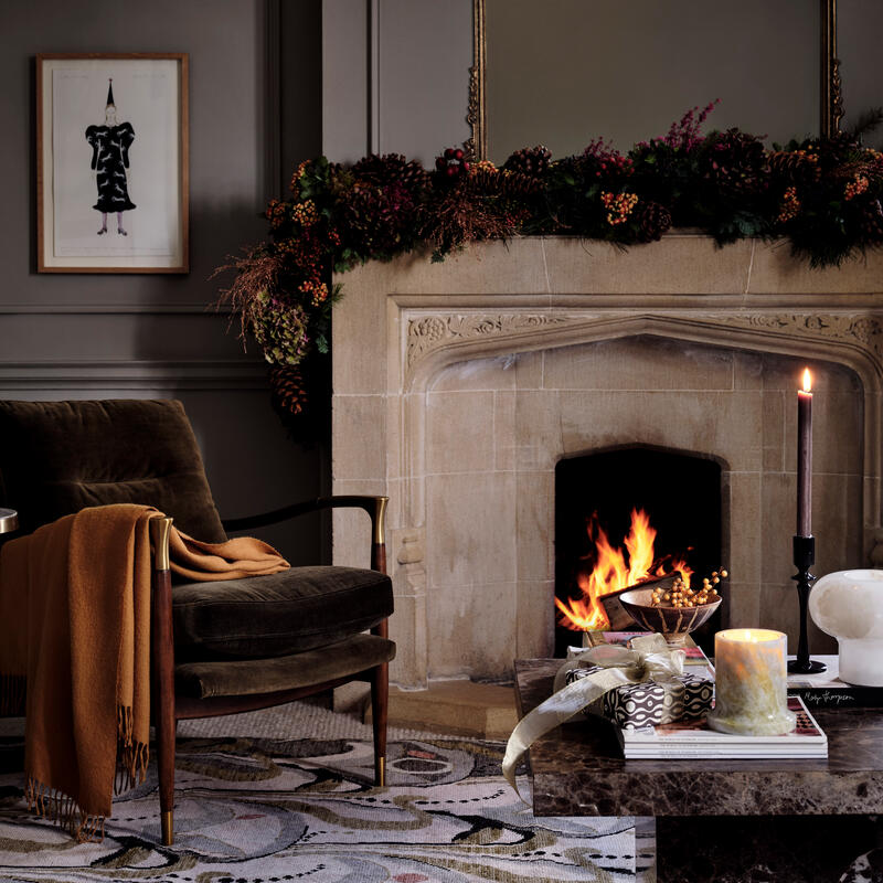 A cozy armchair with an orange blanket sits beside a small table with a wrapped gift and drinks, in a room featuring a lit fireplace adorned with a pinecone garland.