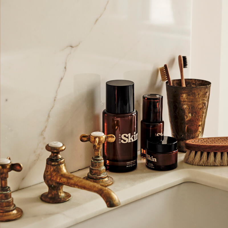 Skin care products with amber bottles labeled "SOHO Skin" placed on a marble bathroom sink counter with vintage brass faucets, alongside a toothbrush in a brass cup and a wooden brush.