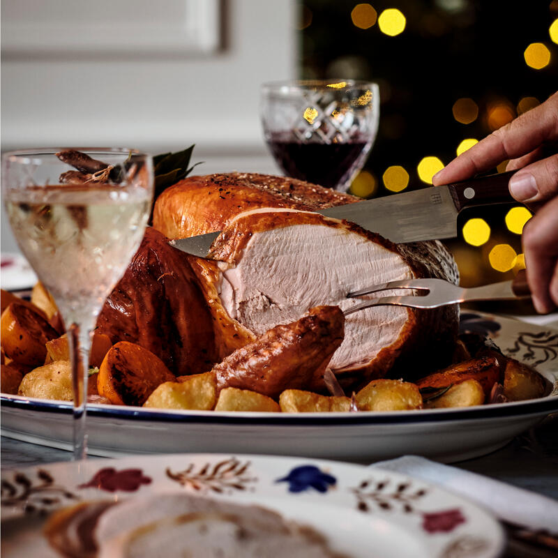 Roast turkey being carved with vegetables on a platter, surrounded by wine glasses on a decorated table; out-of-focus holiday lights in the background.