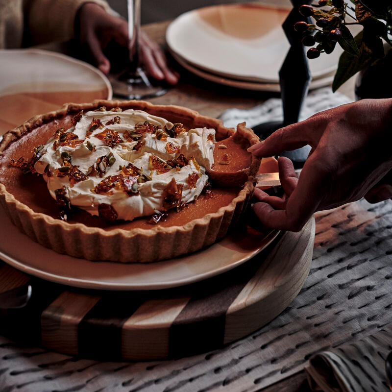 A hand serves a pie topped with whipped cream and nuts at a candlelit dinner table set with plates, utensils, and a glass of red wine.