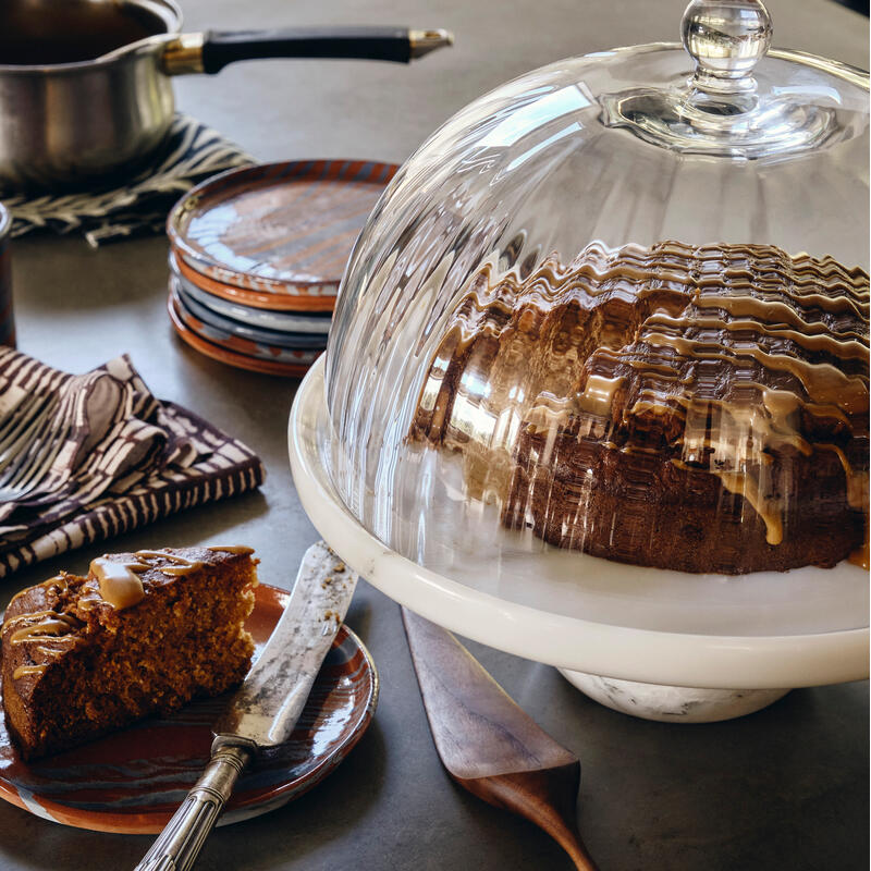 A sliced cake with caramel drizzle is under a glass dome on a table with utensils, napkins, a pot, stacked plates, and a cup, creating a rustic kitchen setting.