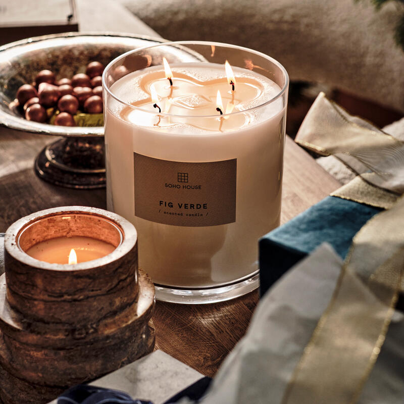 A large glass jar candle labeled "SOHO HOUSE FIG VERDE scented candle" burns on a table next to a smaller lit candle, a bowl of chocolates, and wrapped presents within a cozy, festive setting.