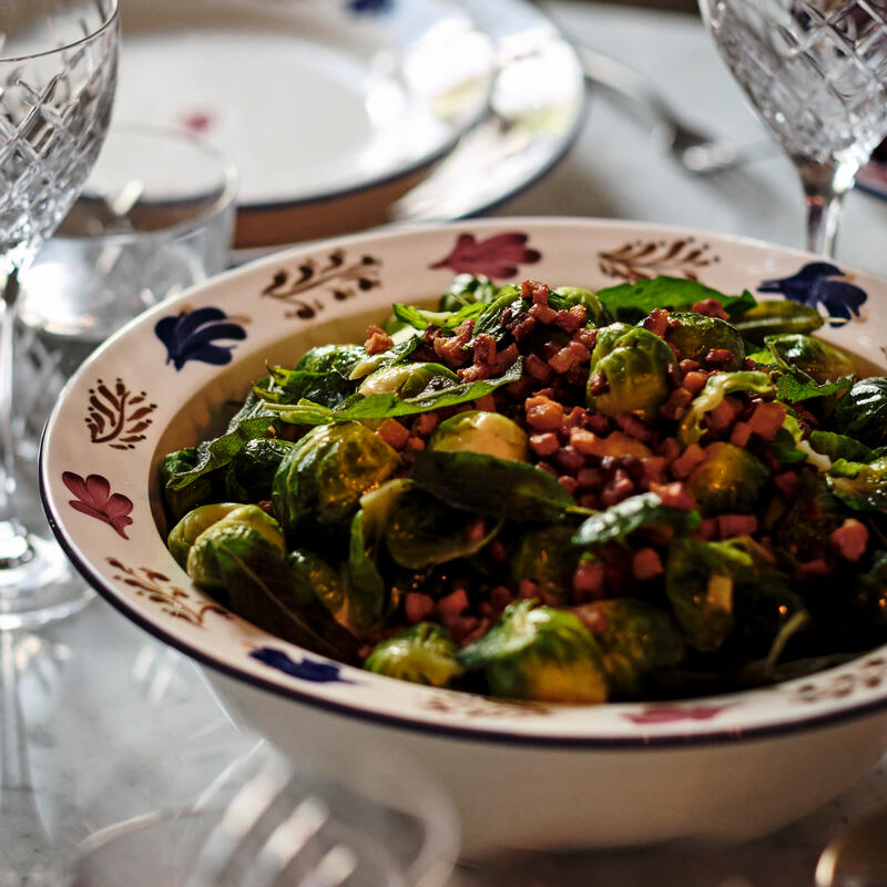 A bowl of Brussels sprouts and diced bacon sits on a table with crystal glasses, plates, and utensils in a dining setting with soft lighting.