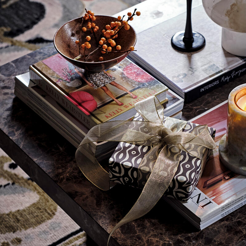 A wrapped gift with a ribbon rests on a stack of books and magazines, accompanied by a decorative bowl of berries and a candle on a marble coffee table in a cozy room.