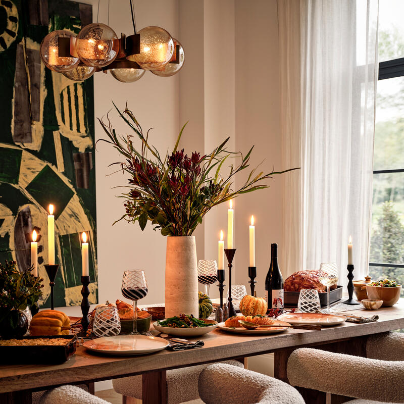 A wooden dining table set for a meal features plates, candles, wine glasses, and food. Plush white chairs surround the table. A large green abstract painting and pendant light hang nearby.