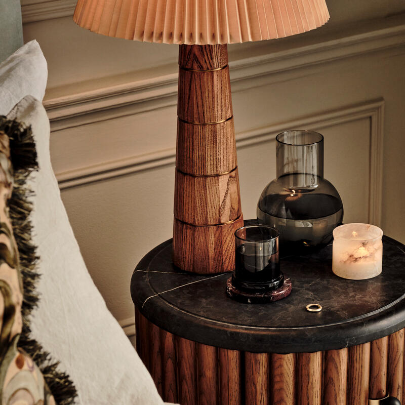 Wooden bedside table holding a large lamp with a pleated beige shade, a candle, a glass vase, and a drinking glass. It sits next to a cream-colored bed, against a paneled wall.