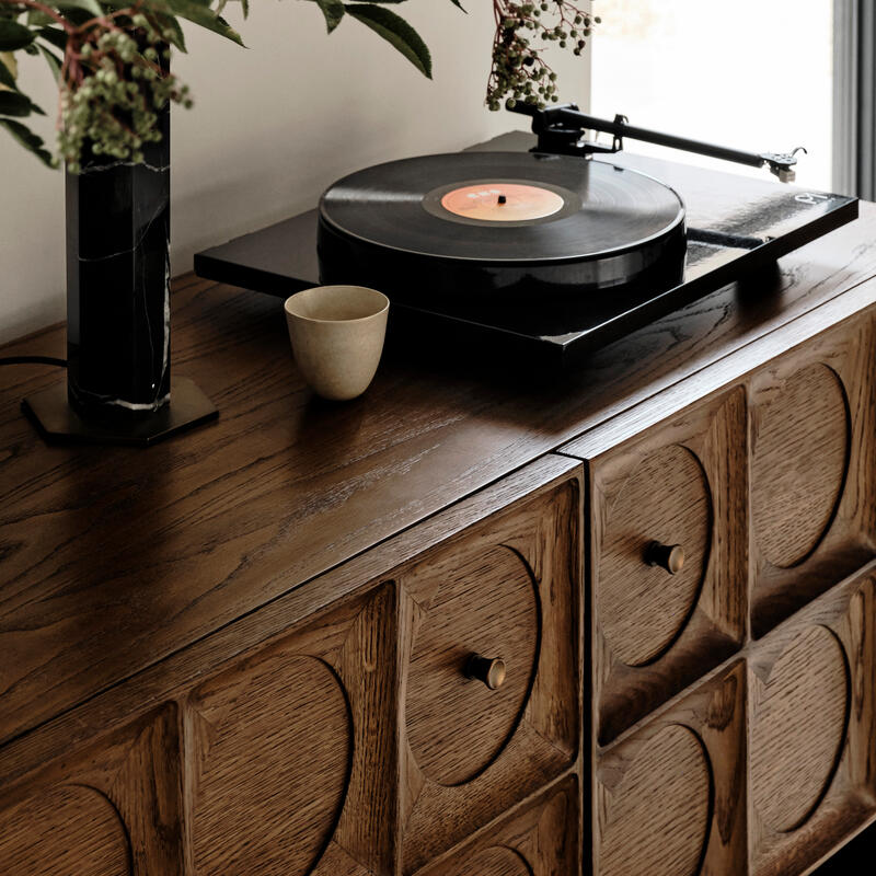 A record player spins a vinyl record atop a mid-century style wooden cabinet. Nearby, a small beige cup and a vase with leafy branches add a touch of nature.