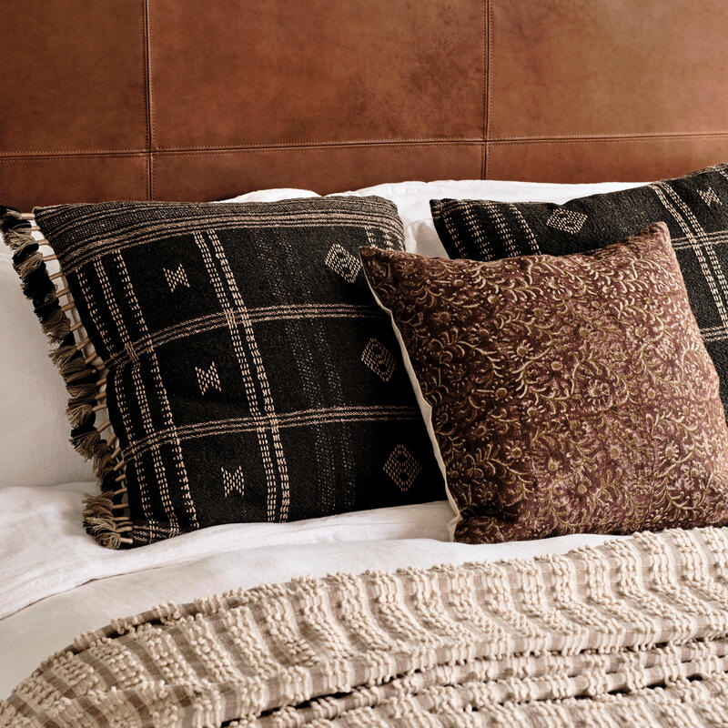 Two decorative pillows rest against a brown leather headboard, one black with geometric patterns and the other brown with floral designs, atop a bed with a white and textured beige blanket.