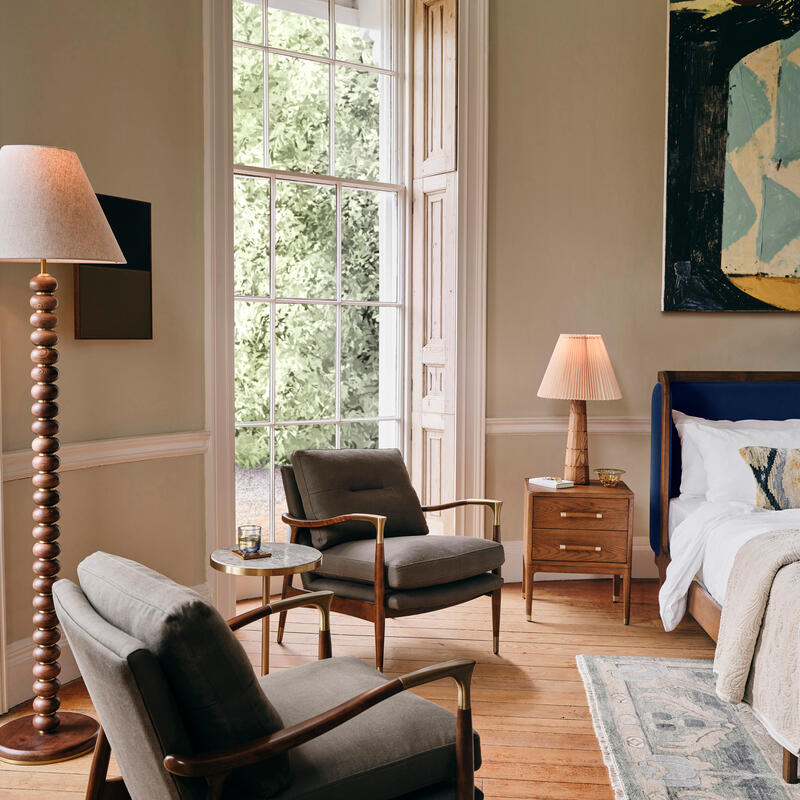 Two mid-century modern armchairs rest on a wooden floor in a cozy bedroom. A large window reveals greenery outside; a bedside table with a lamp and artwork adorn the wall.