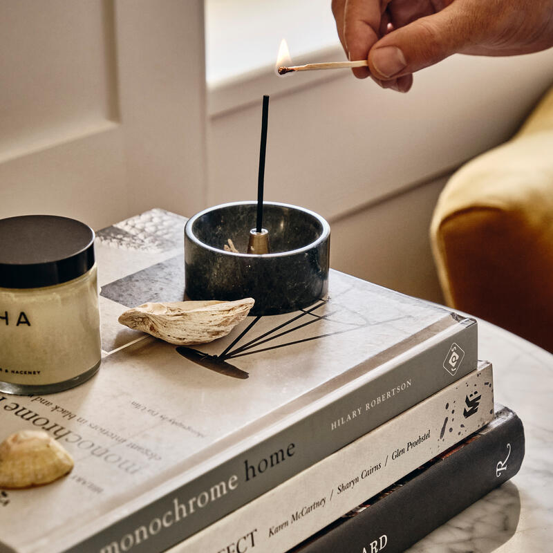 A hand lights an incense stick with a match over a dark stone holder, situated on top of three books titled "monochrome home," "PERFECT IMPERFECT," and "FRANÇOIS HALARD," near a candle.