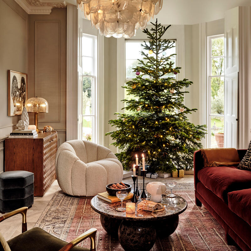 Christmas tree adorned with lights and ornaments, situated beside a bay window; surrounded by cozy furniture, a round coffee table with decor items, a chandelier overhead, and warm ambient lighting.