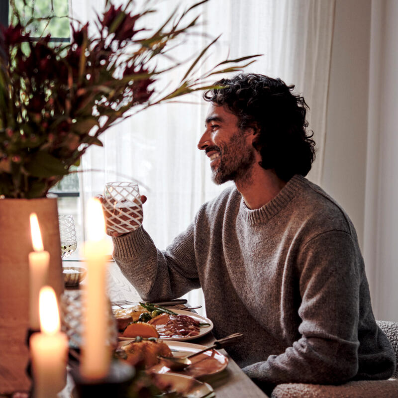 A man, smiling and holding a glass, is sitting at a candlelit dining table with food, in a cozy room adorned with curtains and a vase of flowers.