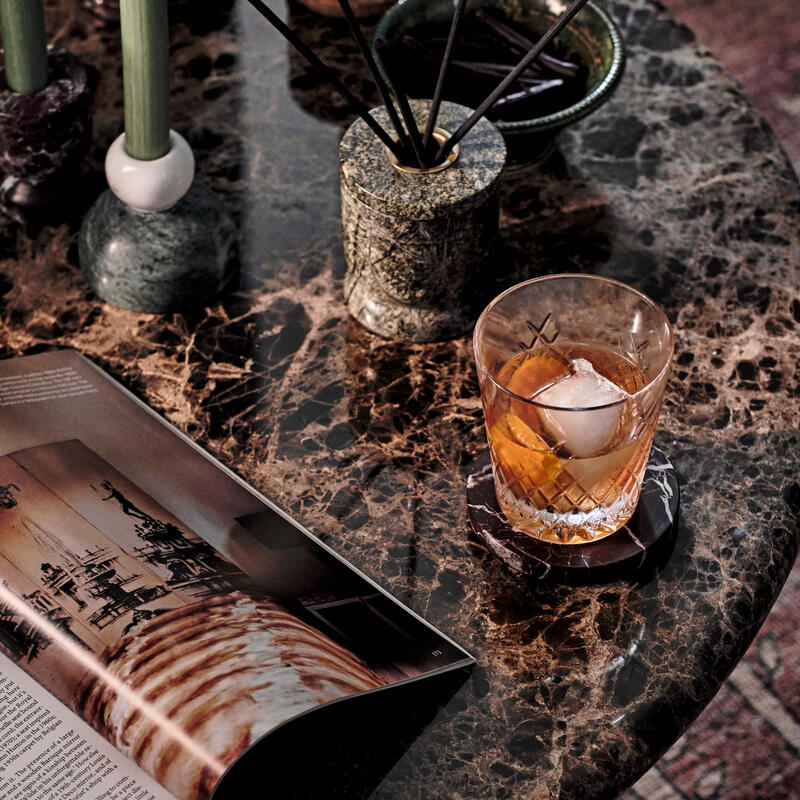 A marble-topped table displays an open magazine, an etched glass of amber liquid with ice, green candles, and a reed diffuser, all casting reflections in a cozy, elegant setting.