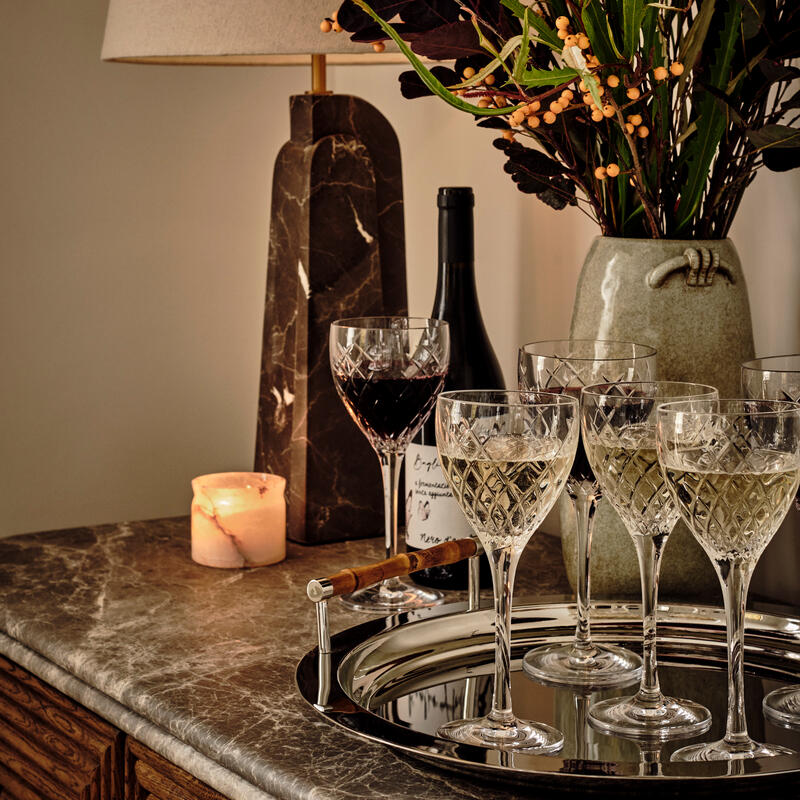 Crystal glasses sit on a silver tray, adjacent to a lit candle, a wine bottle, a vase with foliage, and a marble lamp on a marbled surface in a cozy setting.