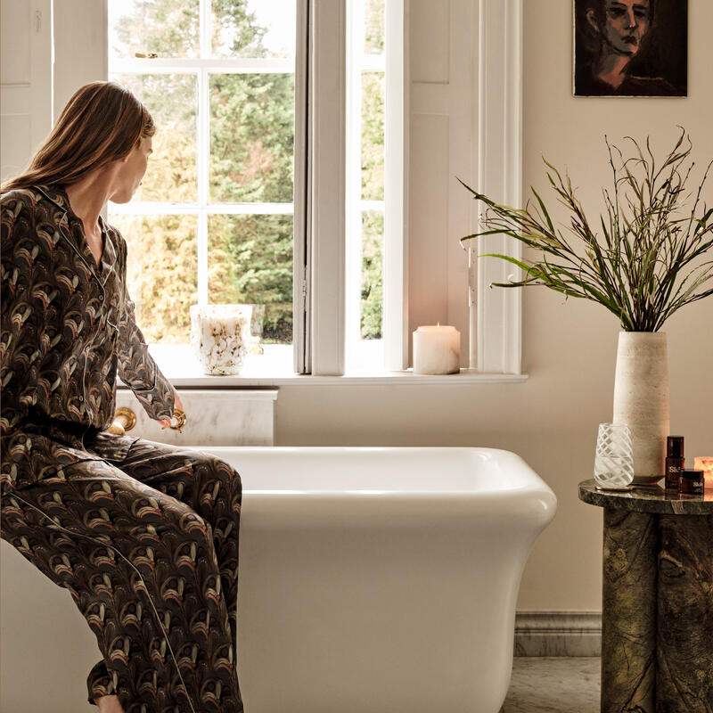 A woman in patterned pajamas sits on a white bathtub, looking out a large window. The bathroom features a marble floor, candles, a potted plant, and a painting on the wall.