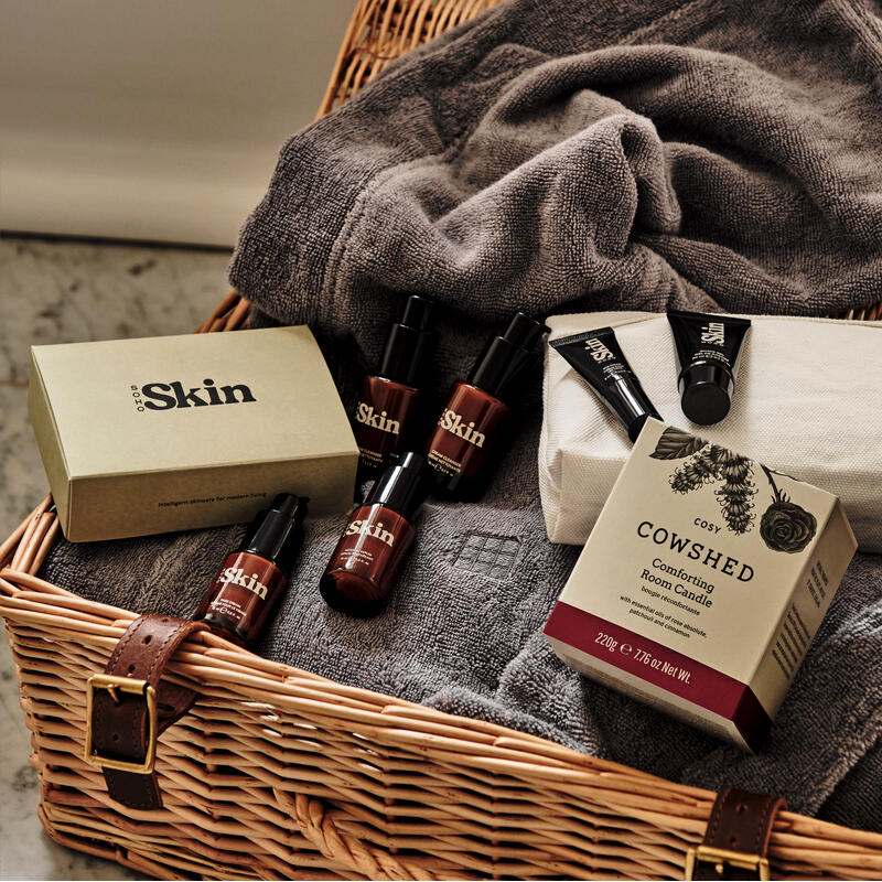 Skincare products lie atop a gray towel inside a woven basket with a marble background. The products include bottles labeled "Pro Skin" and a box labeled "Cowshed Cosy Comforting Room Candle".