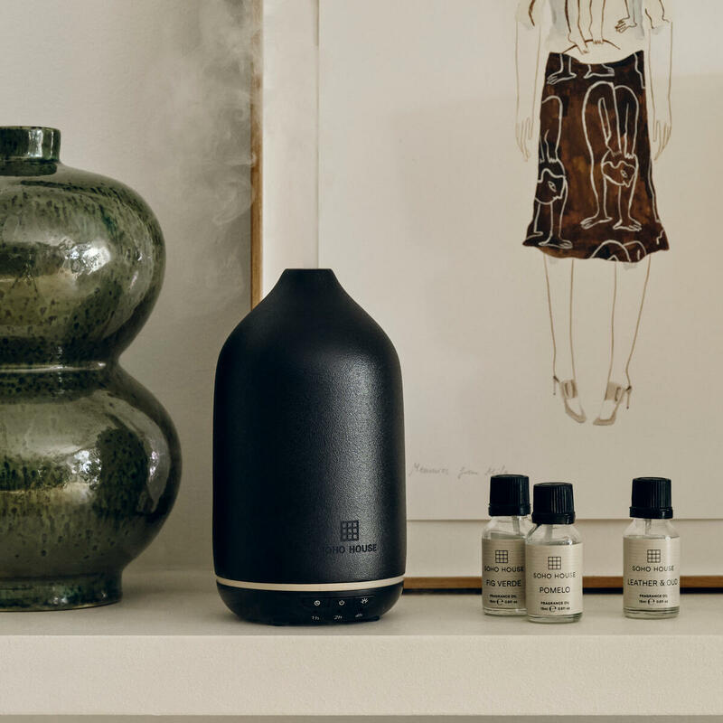 A black SOHO HOUSE essential oil diffuser emitting mist, placed on a white shelf next to a green ceramic vase and three labeled bottles. Behind is a framed minimalist artwork of a standing figure.