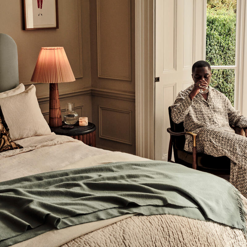 A person sits in a chair drinking from a glass next to a bed with a green blanket, a lamp, and framed artwork in a serene bedroom with a view of greenery through a window.