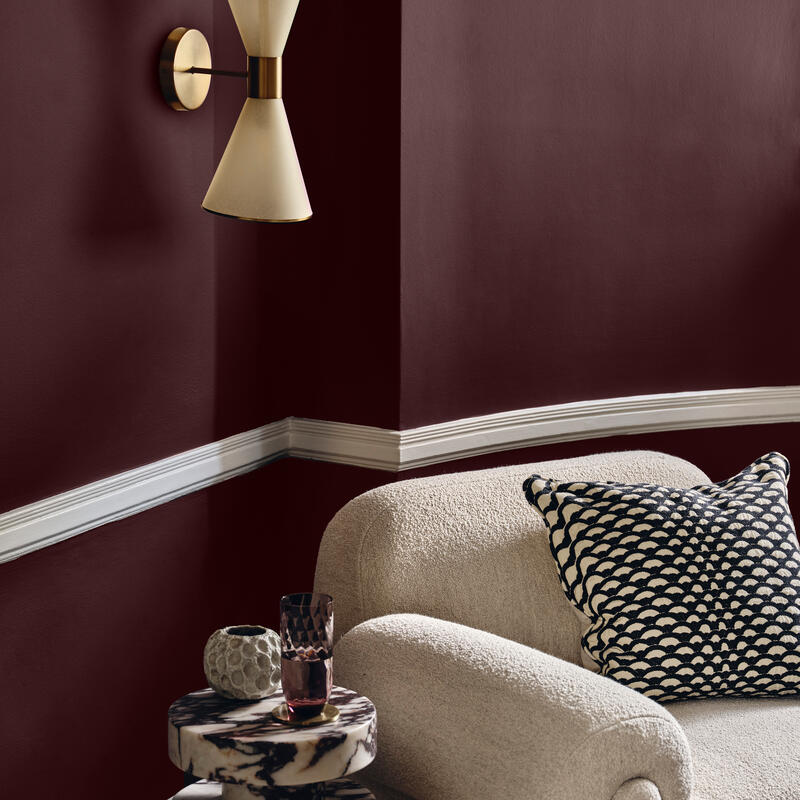 A beige armchair with a patterned pillow sits next to a small marble table holding candles and a glass, beneath a modern wall sconce, in a room with mauve walls and white trim.