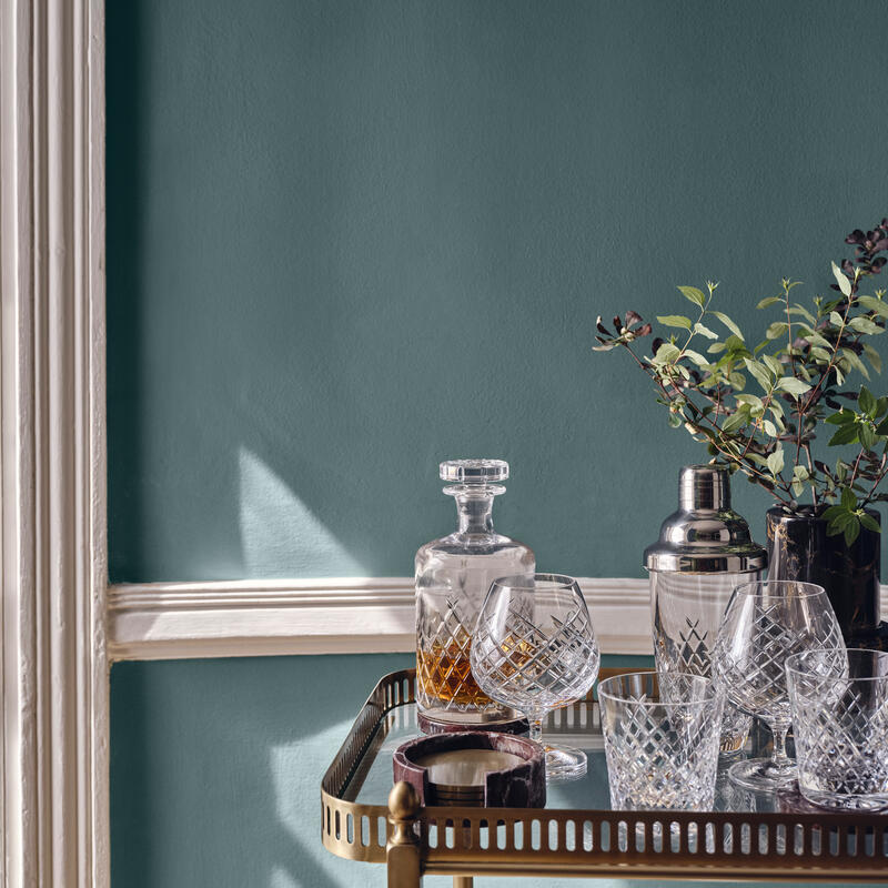 Glass decanter and crystal glasses resting on a brass bar cart with a cocktail shaker, plants, and a teal wall with white trim in the background.