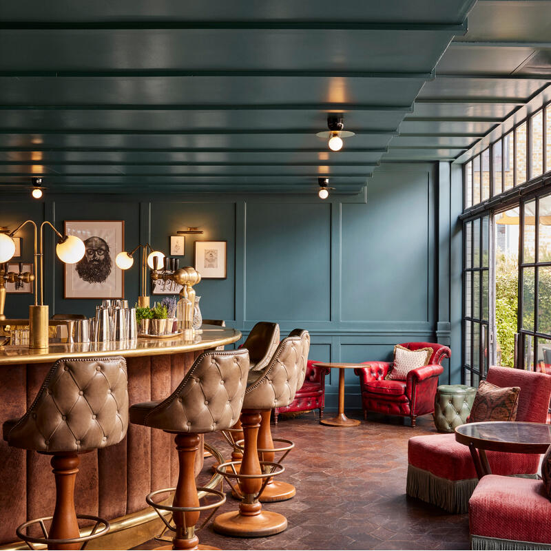 A bar counter with four leather stools fronts a teal wall adorned with framed artwork; red armchairs and small side tables complement the cozy, softly lit, luxurious interior.