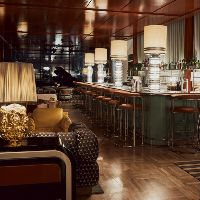 Elegant bar with modern lamps on the counter, brown leather barstools, wooden parquet floor, and cozy seating areas, including a yellow armchair and dark loungers, in a dimly lit, stylish lounge.