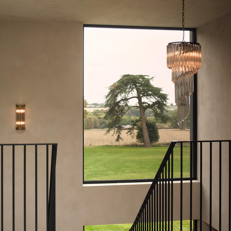 A black metal staircase descends within a modern, beige room featuring a large window framing a verdant landscape. A tiered chandelier and wall sconce provide lighting.