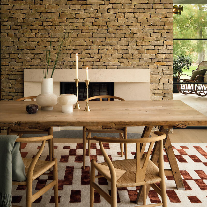 Wooden dining table with candle holders and vases, set on a rug with red patterns, in front of a stone wall with a fireplace; large windows and pendant lights in the background.