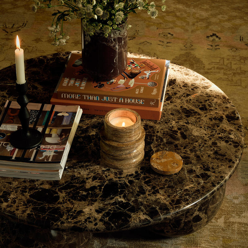 A dark marble tabletop holds a vase of flowers, stacked books titled "More Than Just A House," a lit candle, and a candle holder with a burning candle, in a cozy indoor setting.