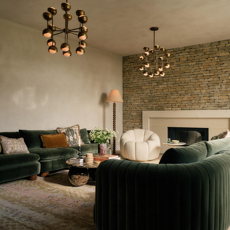 Green velvet sectional sofa forms a cozy seating area around a low coffee table with books and flowers, in a living room with stone fireplace, warm lighting, and wooden floor.