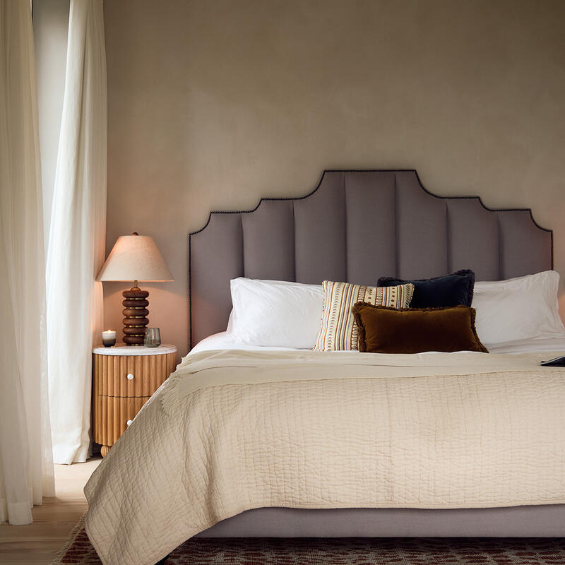 A neatly made bed with a geometric headboard sits between two wooden nightstands with table lamps in a cozy, softly lit bedroom with beige walls, white curtains, and a red patterned rug.