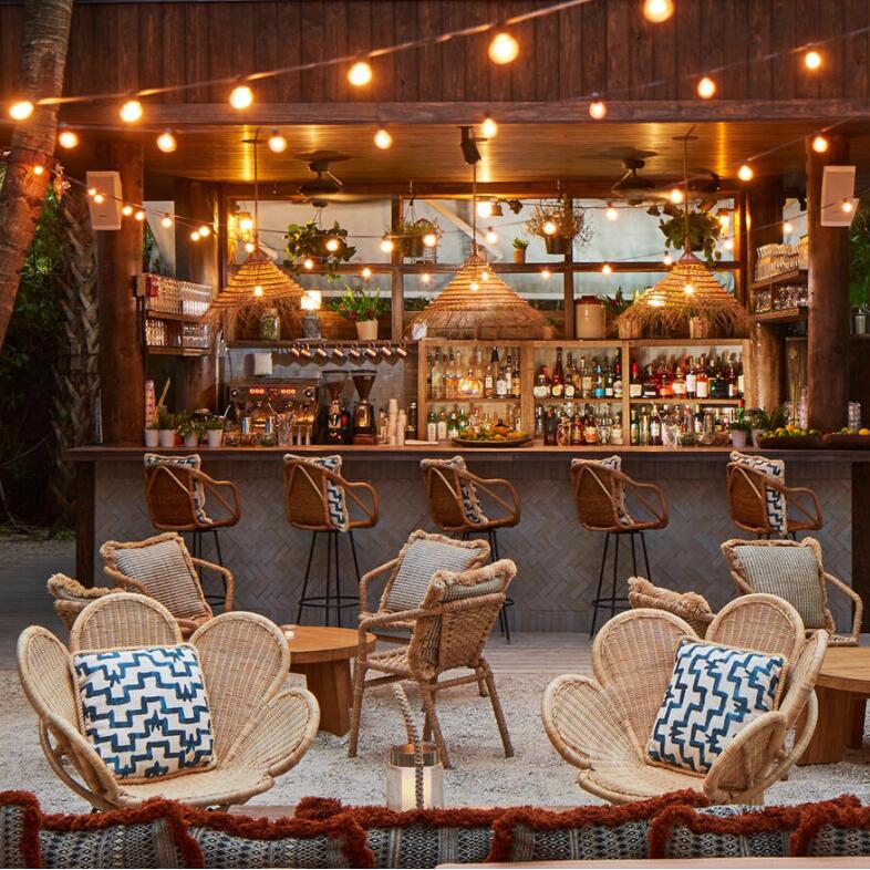 Rattan chairs sit around wooden tables with blue patterned cushions in front of a rustic outdoor bar, adorned with hanging lights and plants, surrounded by a leafy tropical setting.