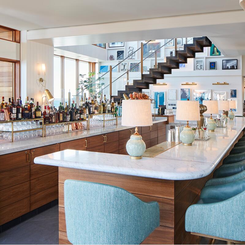 A chic, modern bar featuring a marble countertop lined with bottles and glasses, with teal chairs and table lamps, situated in a bright, airy room with an ocean view and stylish decor.