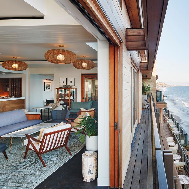 Modern living room with wooden furniture and woven light fixtures opens onto a balcony overlooking a sandy beach and expansive ocean with gentle waves.