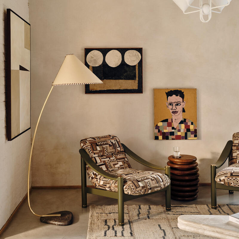 Two patterned armchairs with wooden side table holding a glass, next to a floor lamp, in a modern living room featuring abstract wall art, a portrait, and a light fixture hanging from the ceiling.