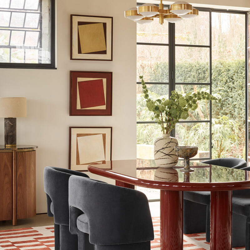 A modern dining area with a dark red oval table, six black chairs, a large plant centerpiece, framed abstract art, and large windows leading to a garden, under a rustic wooden ceiling.