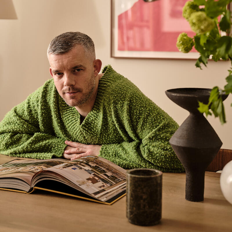 A person in a green sweater leans on a wooden table, looking at an open book. The setting includes a framed portrait, a black vase, and green plant.