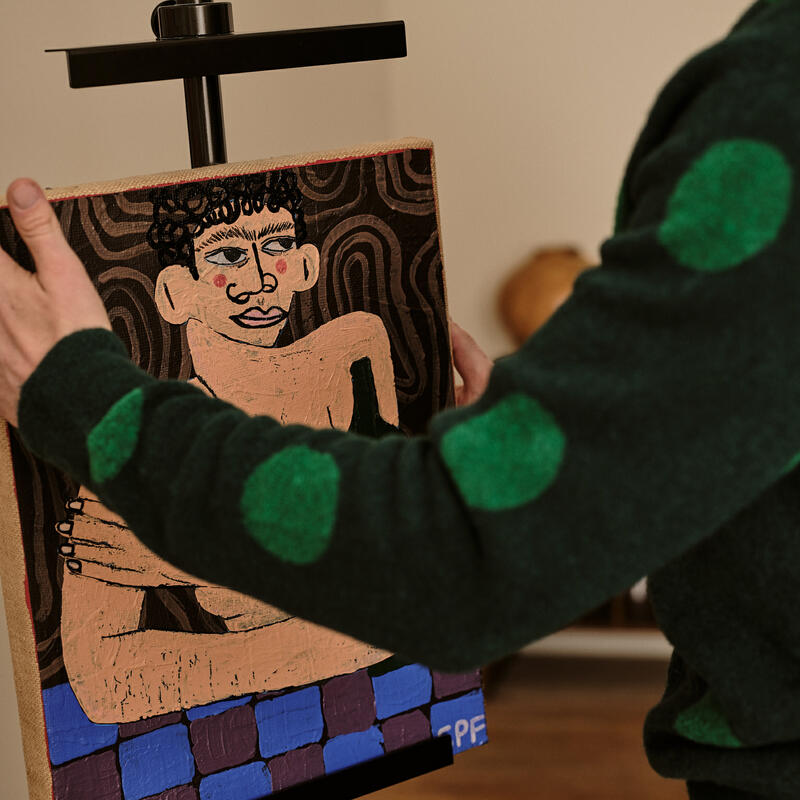 A person in a green polka-dot sweater arranges a colorful painting of a stylized human figure on an easel in a well-lit room with hardwood floors.