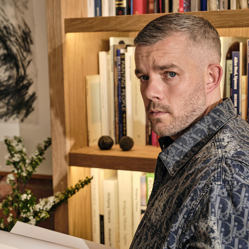 A man in a patterned shirt stands reading a book in front of a wooden bookshelf filled with books and a small sculpture, with green floral arrangements and artwork in the background.