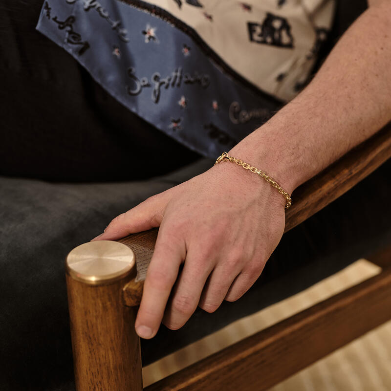 A hand with a gold chain bracelet rests on the wooden arm of a black cushioned chair. The person is wearing a shirt adorned with zodiac signs and names like "Sagittarius."