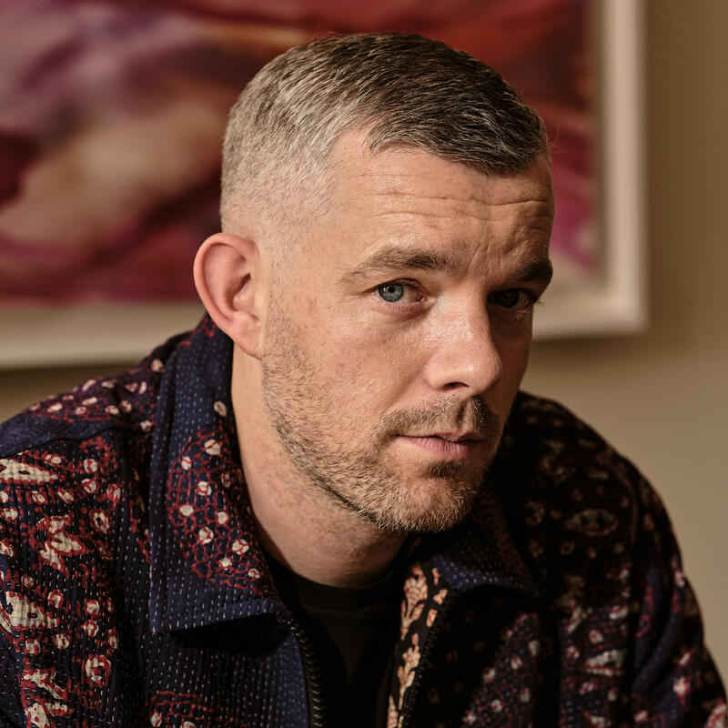 A man with short hair and a beard appears thoughtful, wearing a patterned jacket, indoors against a colorful, abstract painting background.