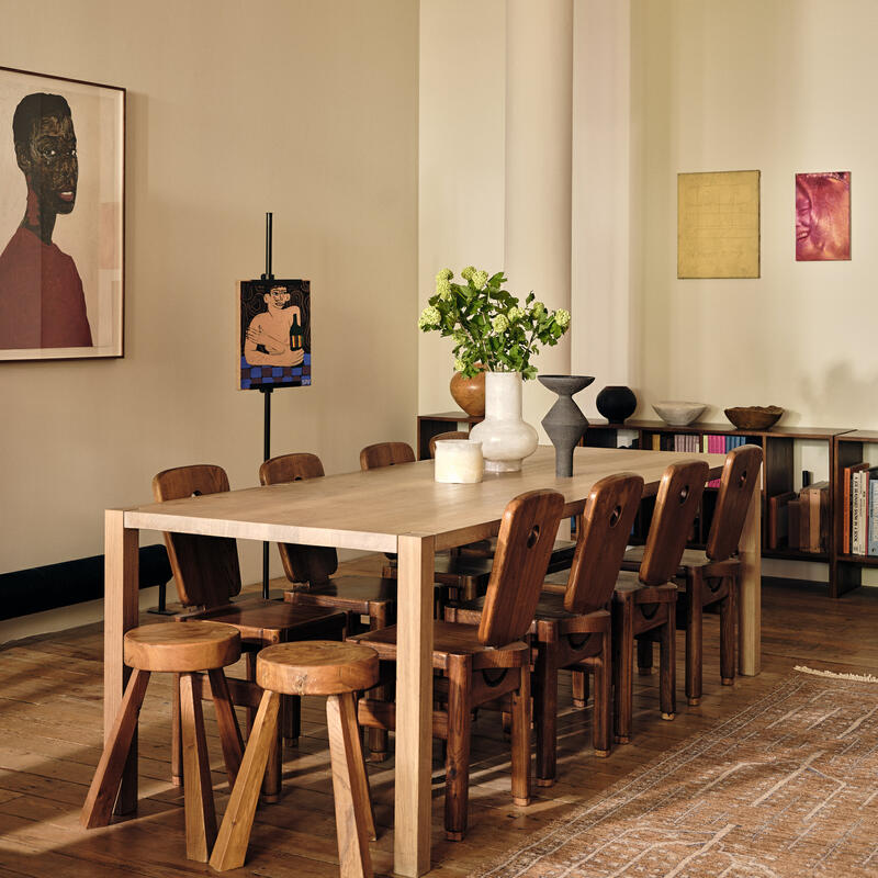 A wooden dining table surrounded by chairs holds vases with flowers, situated in a room with art on the walls, a large column, and a bookshelf filled with books.