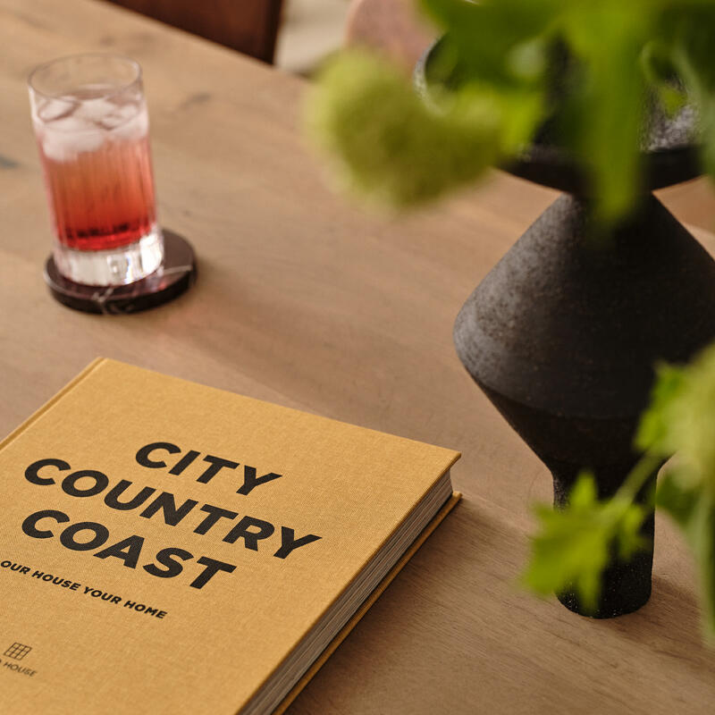 A hardcover book titled "City Country Coast" lies on a wooden table beside a glass of red beverage, a black vase with green flowers, and a wooden chair in a cozy interior setting.