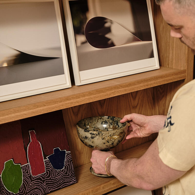 A person places a decorative bowl on a wooden shelf, which also holds framed photographs and colorful artwork.