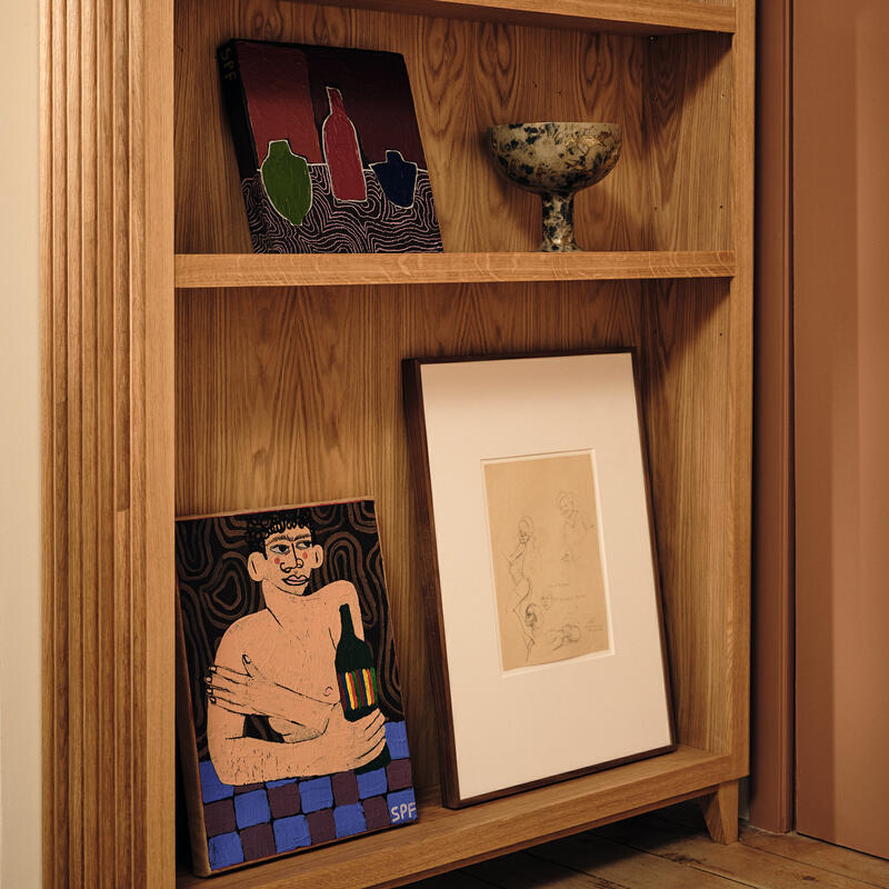 Wooden bookshelf displaying various art pieces, including framed drawings, a colorful painting of a figure holding a bottle, pottery items, and a figurine, in a softly lit room.