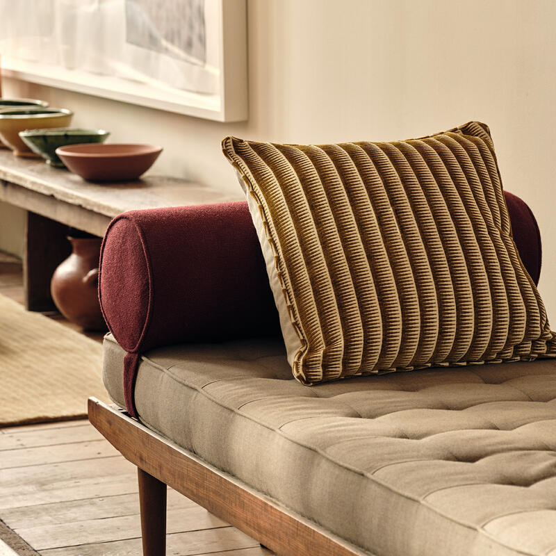 A beige cushioned daybed featuring a striped pillow and red bolster sits in a cozy, earthy-toned room with pottery and framed artwork on a wall-mounted shelf.