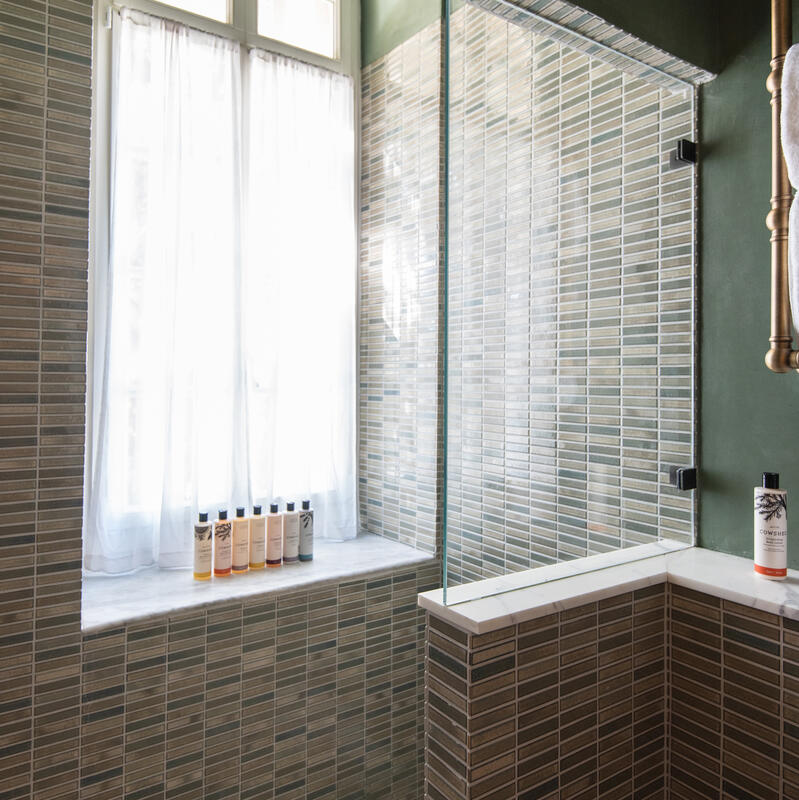 Shower stall with green tiled walls and white tiled floor, showcasing several bottles of toiletries on a window ledge with sheer curtains adjacent to a glass partition and brass fixtures.