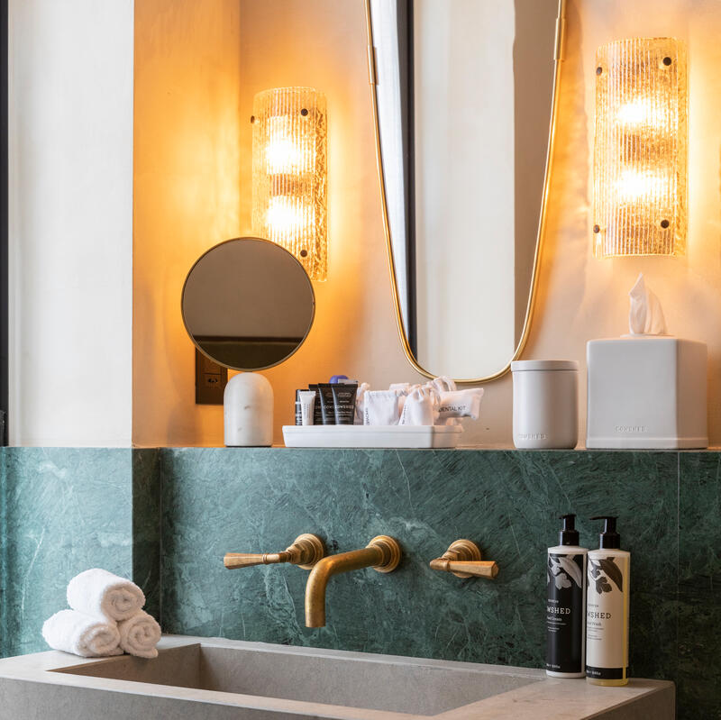 A modern bathroom sink with gold faucets under a mirror between illuminated wall sconces, surrounded by green marble, features towels, toiletries, and a tissue box on the counter. Text on the boxes reads, "COWSHED."